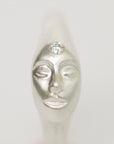A silver sculpted ring shaped like a stylized face with closed eyes and a clear round gemstone set on the forehead, shown against a plain light gray background.