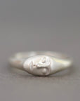 A sterling silver ring featuring a stylized, minimalist face with closed eyes, set against a soft, out-of-focus gray background.
