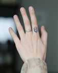 A close-up of a person's hand showing two rings: a silver band ring with a small clear stone on the ring finger, and a small dark geometric tattoo or symbol on the middle finger.