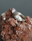 A silver ring with a stylized face engraving and a small clear stone, displayed on a textured brown crystal cluster against a dark gray background.
