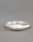 A sterling silver ring featuring a sculpted face with closed eyes and a small round clear gemstone on its cheek, set against a neutral gray background.