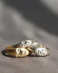 Three sculptural face-shaped rings in polished yellow gold, silver, and white gold, stacked together on a textured gray surface with soft shadows.