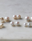 Six pairs of pearl studs in three sizes and two metals, photographed on white marble. The back row shows, left to right, the large pearl studs with gold barnacles, medium pearl studs with gold barnacles, and large pearl studs with silver barnacles. The front row shows, left to right, the little pearl studs with gold barnacles, the medium pearl studs with silver barnacles, and the little pearl studs with silver barnacles.