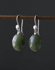 A set of earrings featuring silver or gold scallop shells attached to green jade spheres, displayed on a dark background.