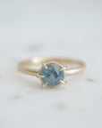 a muted teal round sapphire sits within gold prongs in this gold ring, a hidden halo of diamonds beneath. Front view.