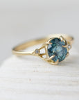 oval teal montana sapphire is set in a gold branch ring with vine-like prongs holding the stone in. Three little diamond berries adorn the branches. 1/4 view.