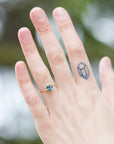 oval teal montana sapphire is set in a gold branch ring with vine-like prongs holding the stone in. Three little diamond berries adorn the branches. Photographed on hand.