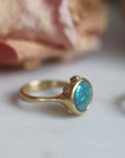 Gold ring with a blue opal on a light background with flowers in the background