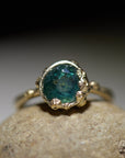 a deep teal montana sapphire is set in a gold bezel of a gold ring, with barnacles coming up over the side of the bezel and onto the stone, as well as along the band. front view.