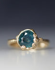 a deep teal montana sapphire is set in a gold bezel of a gold ring, with barnacles coming up over the side of the bezel and onto the stone, as well as along the band. front view.