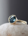 a deep teal montana sapphire is set in a gold bezel of a gold ring, with barnacles coming up over the side of the bezel and onto the stone, as well as along the band. 1/4 view.