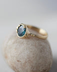 a deep teal montana sapphire is set in a gold bezel of a gold ring, with barnacles coming up over the side of the bezel and onto the stone, as well as along the band. side view.