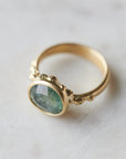 aqua colored teal montana sapphire stone set in a bezel setting of a gold ring with barnacles in a nautical aesthetic. Photographed at 1/4 view, with a marble background.