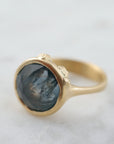 steely blue color change sapphire set in a gold bezel and band. Around the cup of the bezel are tiny gold barnacles. Photographed side view and in natural light.