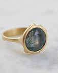 steely blue color change sapphire set in a gold bezel and band. Around the cup of the bezel are tiny gold barnacles. Photographed side view and in natural light.
