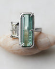 long, rectangular tourmaline stone set with prongs next to a diamond baguette. The ring is platinum, and shown balancing on a shell.
