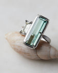 long, rectangular tourmaline stone set with prongs next to a diamond baguette. The ring is platinum, and shown balancing on a shell.