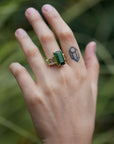 a grand center rectangular green tourmaline gemstone is set within four prongs. On one side is one diamond baguette, on the other sit two. The four stones make an off-set centerpiece of a gold ring. Shown on hand.