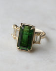 a grand center rectangular green tourmaline gemstone is set within four prongs. On one side is one diamond baguette, on the other sit two. The four stones make an off-set centerpiece of a gold ring.