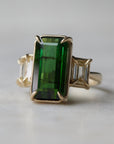 a grand center rectangular green tourmaline gemstone is set within four prongs. On one side is one diamond baguette, on the other sit two. The four stones make an off-set centerpiece of a gold ring.