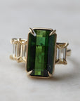 a grand center rectangular green tourmaline gemstone is set within four prongs. On one side is one diamond baguette, on the other sit two. The four stones make an off-set centerpiece of a gold ring.