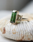 a grand center rectangular green tourmaline gemstone is set within four prongs. On one side is one diamond baguette, on the other sit two. The four stones make an off-set centerpiece of a gold ring. Shown on an ammonite fossil.