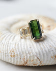 a grand center rectangular green tourmaline gemstone is set within four prongs. On one side is one diamond baguette, on the other sit two. The four stones make an off-set centerpiece of a gold ring. Shown on an ammonite fossil.