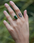 a grand center rectangular green tourmaline gemstone is set within four prongs. On one side is one diamond baguette, on the other sit two. The four stones make an off-set centerpiece of a gold ring. Shown on hand.