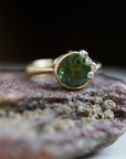 A 2.74ct tourmaline ring. The tourmaline is a deep green and is set in 14k gold with a 14k gold band. The setting is encrusted with bright sterling silver barnacles. The ring is viewed from the front and sits on a stone.