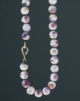 purple and white swirly beads of round quahog shells are strung on black silk and have gold barnacles attached throughout. A gold fish hook clasp attached the strand. Detail shot on dark grey background