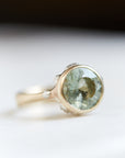 A 2.56ct Aquamarine ring. The ring is set in 14k gold with a tapered 14k gold band. The Aquamarine is a pale green with speckles. The setting is encrusted with bright sterling silver barnacles. The ring is on a white background.