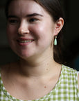 Woman wearing a green checkered dress and green drop earrings with a blurred dark background