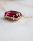 pink rectangular stone with in gold prongs on a gold chain with diamonds set in a hidden halo on the side of the setting. photographed on white marble.
