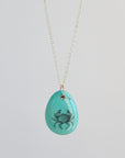 Teal pendant with a black crab design on a silver chain against a light gray background
