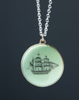 melon green round variscite cabochon is set in a gold bezel and hangs beneath a silver chain. an old whaling ship is hand-etched into the surface with black ink.