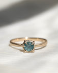 14k yellow gold vanity ring with a round teal blue Montana sapphire and a halo of small diamonds around the setting, on a white fabric background.