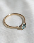 14k yellow gold vanity ring with a round teal blue Montana sapphire and a halo of small diamonds around the setting, on a white fabric background, side view.
