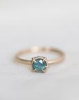 14k yellow gold vanity ring with a round teal blue Montana sapphire and a halo of small diamonds around the setting, on a white fabric background.