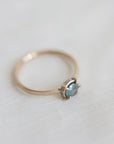 14k yellow gold vanity ring with a round teal blue Montana sapphire and a halo of small diamonds around the setting, on a white fabric background, side view.