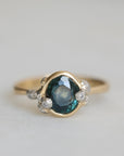 Gold ring with a teal gemstone and diamond barnacle accents on a light gray background