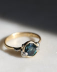 Gold ring with a teal gemstone and diamond barnacle accents on a light gray background