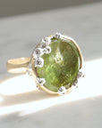a lime green, rough-hewn tourmaline gemstone is set in a light yellow gold. Lots of little silver barnacles wrap around the setting and up over the stone. Photographed in sunlight.