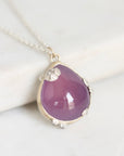 A teardrop-shaped purple gemstone pendant in a silver bezel setting with small shell-like silver accents, hanging from a silver chain on a light marble surface.