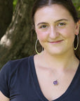 A woman with dark hair wearing a silver necklace with a purple heart pendant and large gold hoop earrings, posing outdoors in front of a tree.