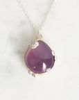 A teardrop-shaped purple gemstone pendant in a gold and silver mixed metal setting, hanging from a delicate silver chain against a light marble background.