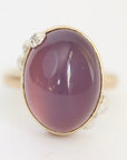 An oval cabochon gemstone ring in a gold setting with small clear accent stones on the band, shown on a plain white background.