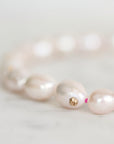 A close-up of a pearl beaded bracelet on a light marble surface. The bracelet features irregular, lustrous pale pink pearls strung with small bright pink beads and a small gold flower-shaped charm in the center.