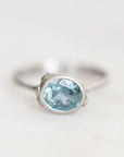 A silver ring featuring an oval-cut light blue gemstone in a bezel setting, photographed on a clean, light off-white background.