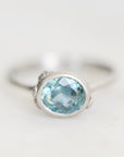 A silver ring featuring an oval-cut light blue gemstone in a bezel setting, photographed on a clean, light off-white background.