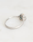 A silver ring with a decorative head featuring small clear stones and a tiny blue-green accent, photographed on a light neutral background.